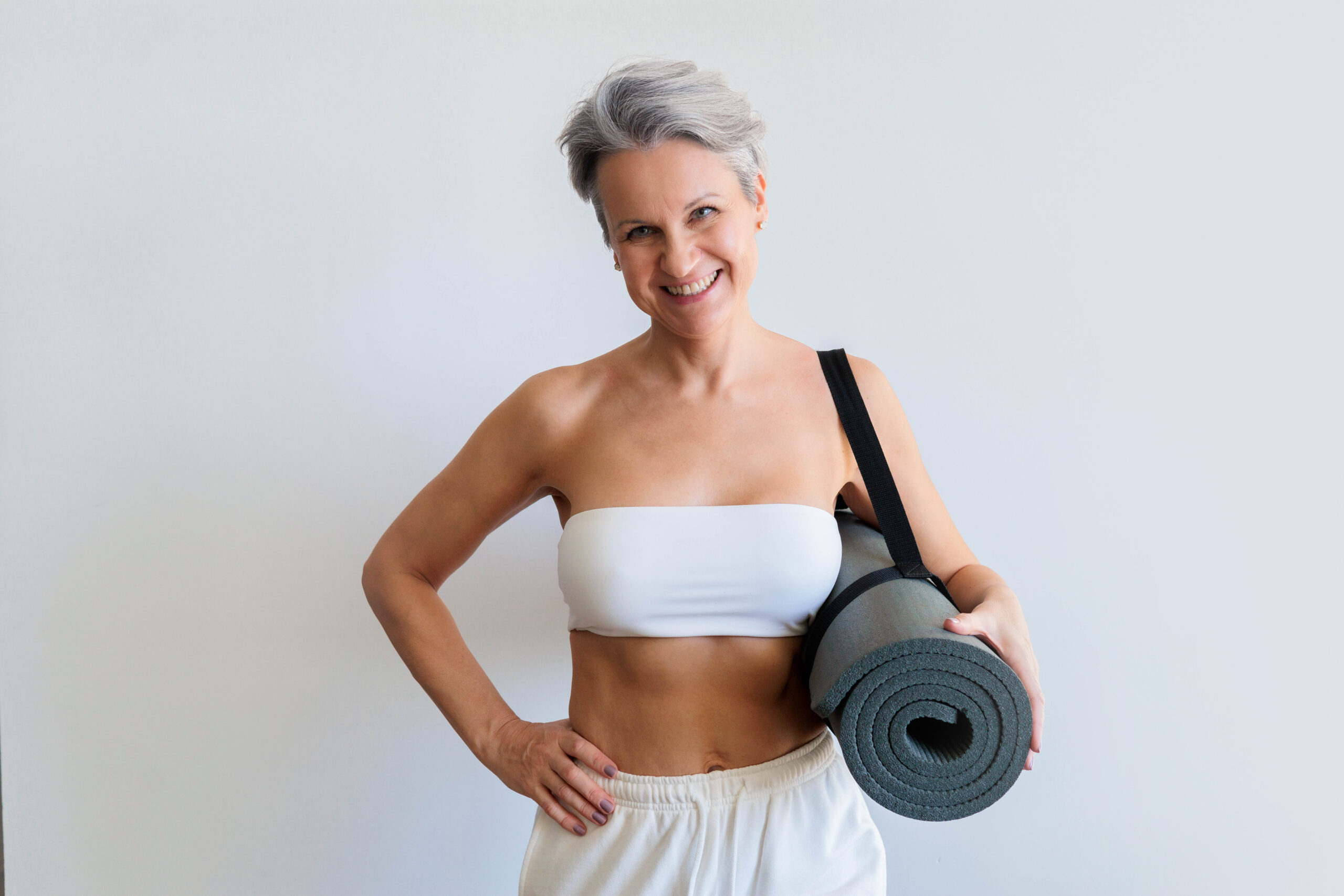 Smiling mature gray haired sport woman with yoga roll mat posing isolated on white background. Happy mid age senior beautiful athletic female enjoy healthy lifestyle gymnastic pilates at copy space